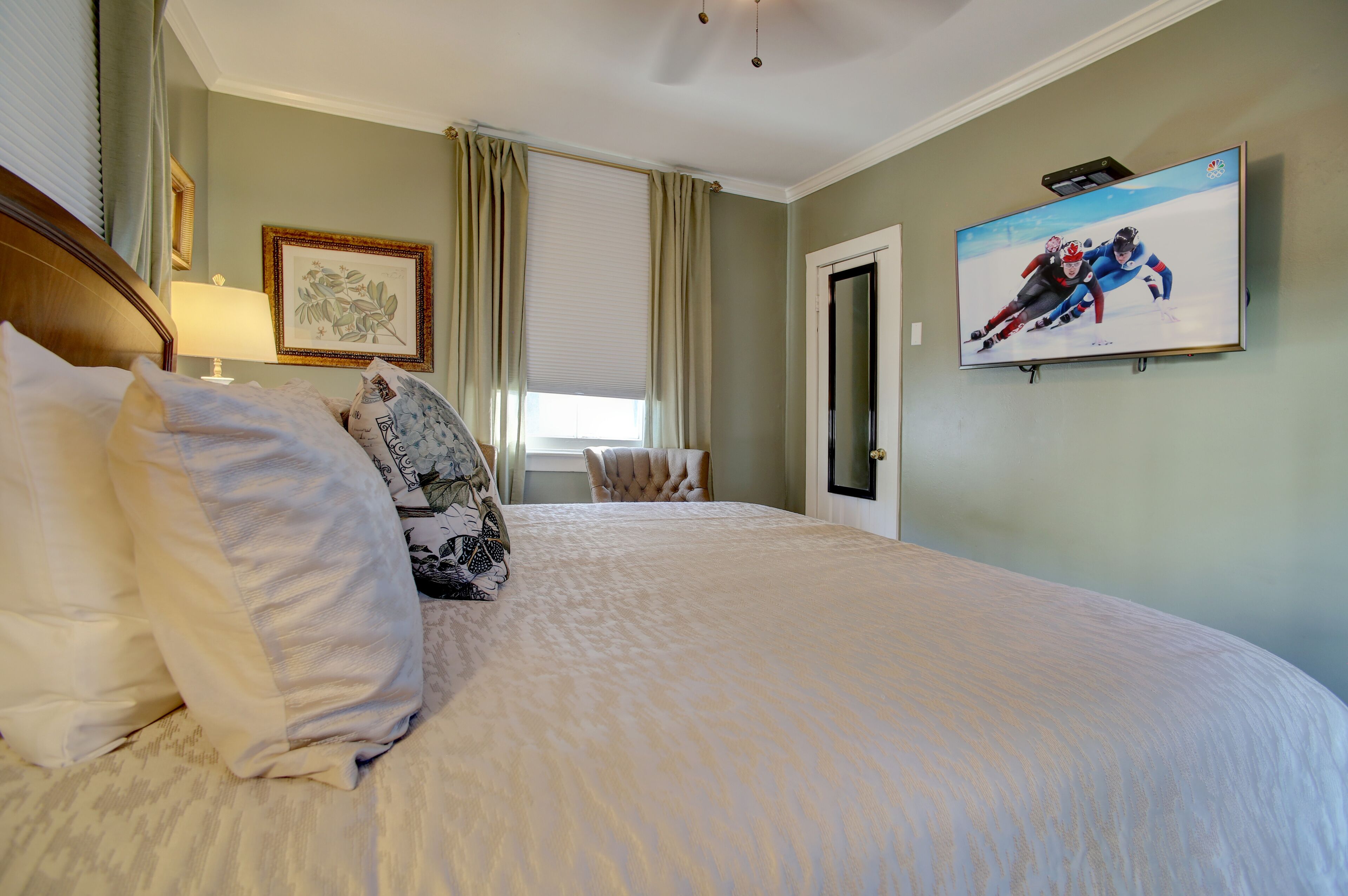 Un dormitorio con una cama grande, un televisor montado en la pared, una ventana con cortinas y un cuadro enmarcado en la pared.