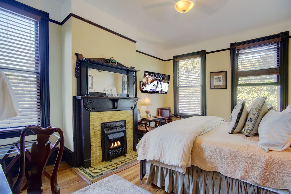 A bedroom with a fireplace, a bed with a beige bedspread, a television mounted on the wall, and a window with blinds.