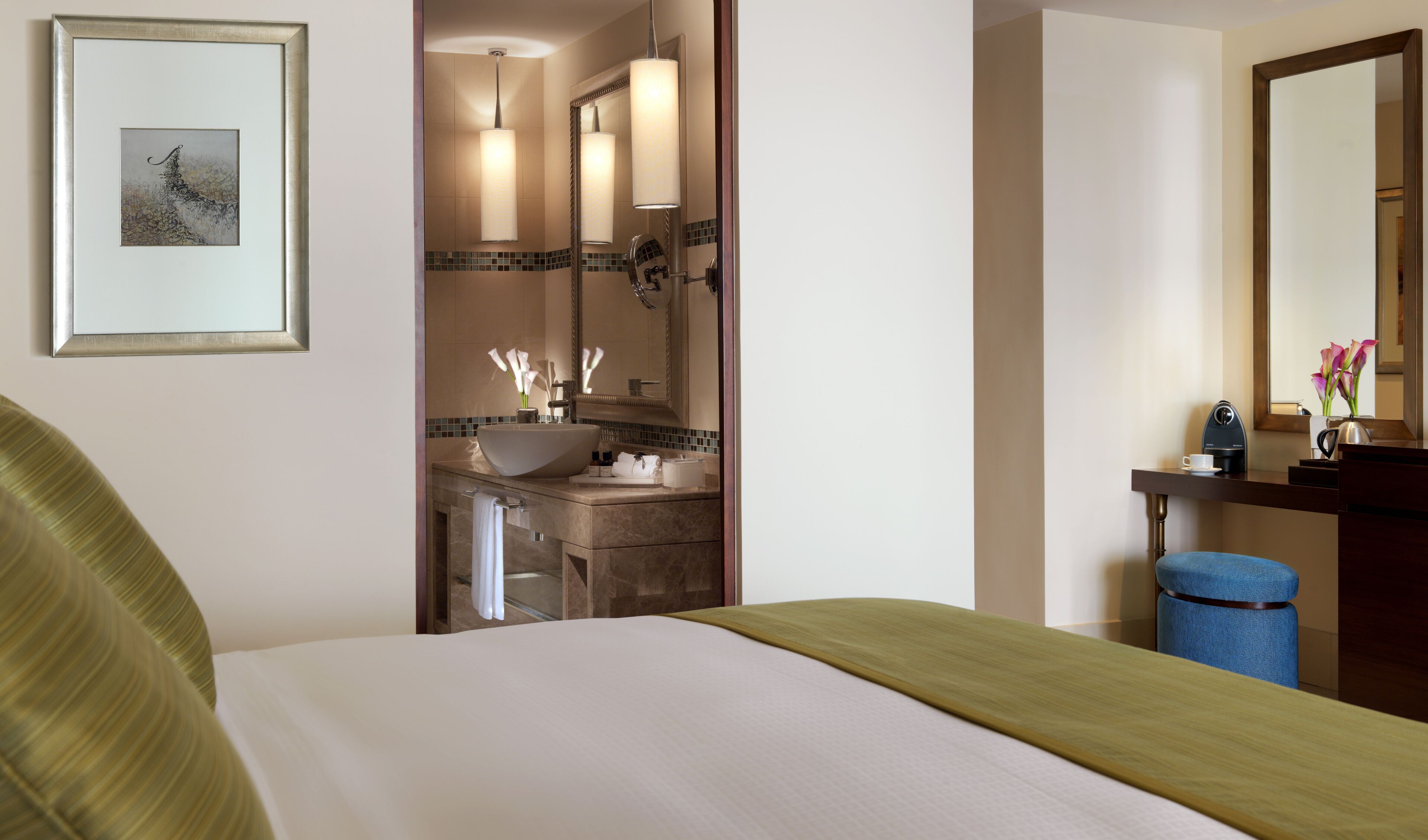 Habitación de hotel con cama, baño con lavabo y espejo, y escritorio con taza y flores.