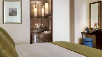 Habitación de hotel con cama, baño con lavabo y espejo, y escritorio con taza y flores.