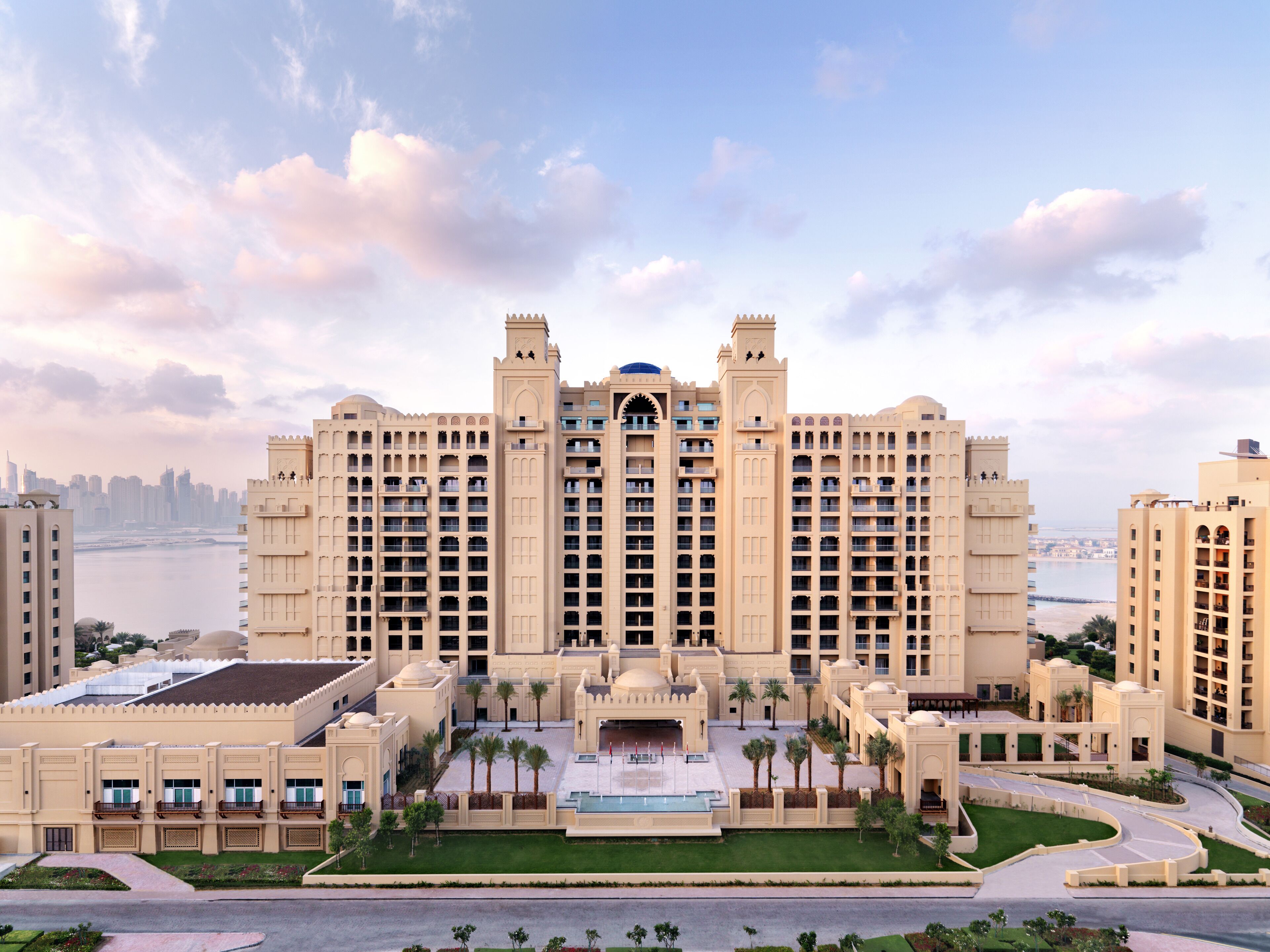 A large, multi-story hotel with a central courtyard, palm trees, and a view of the city skyline.