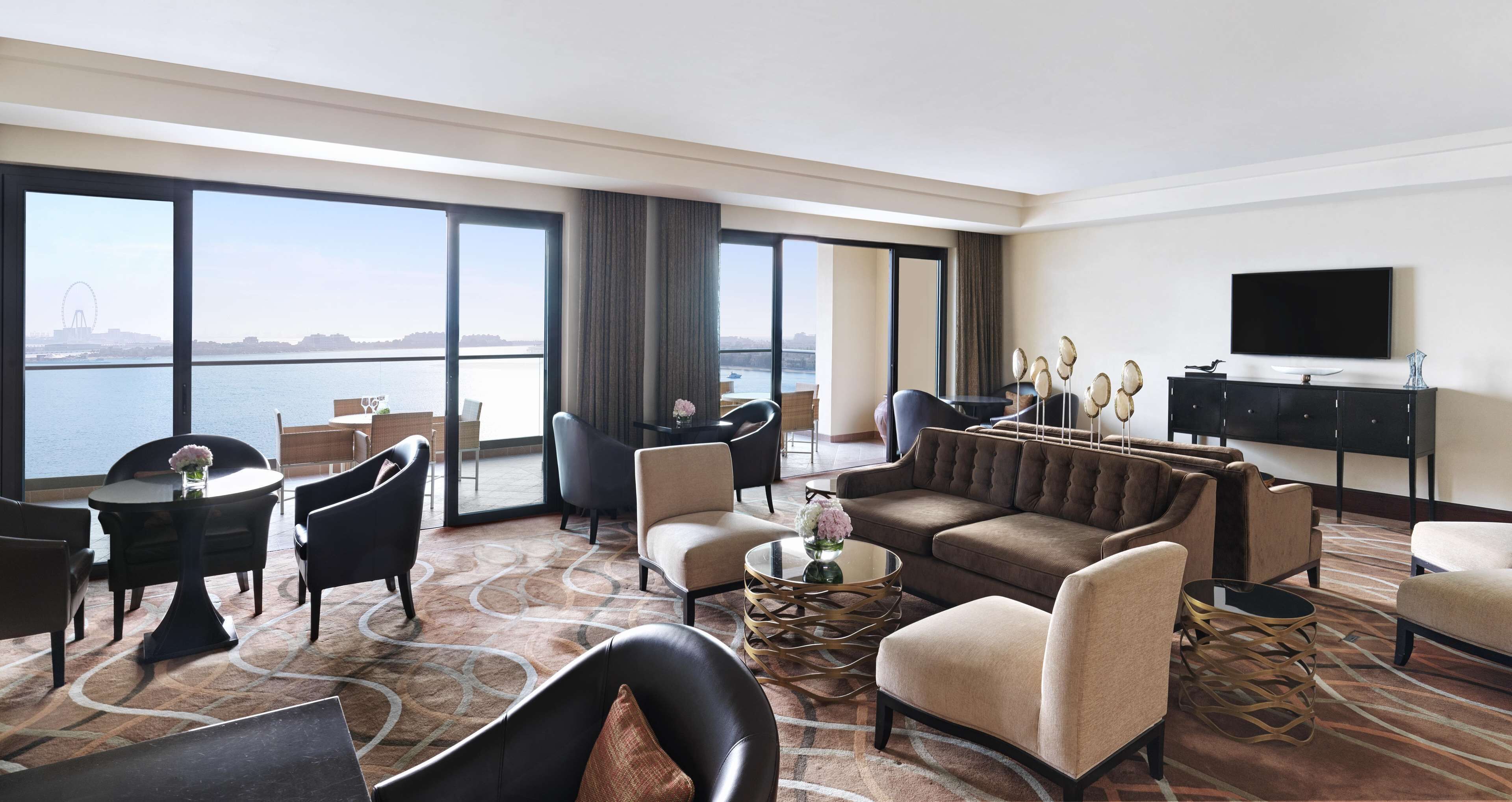 A spacious hotel room with a large window offering a view of a body of water and a bridge. The room is furnished with a sofa, armchairs, a coffee table, and a TV.