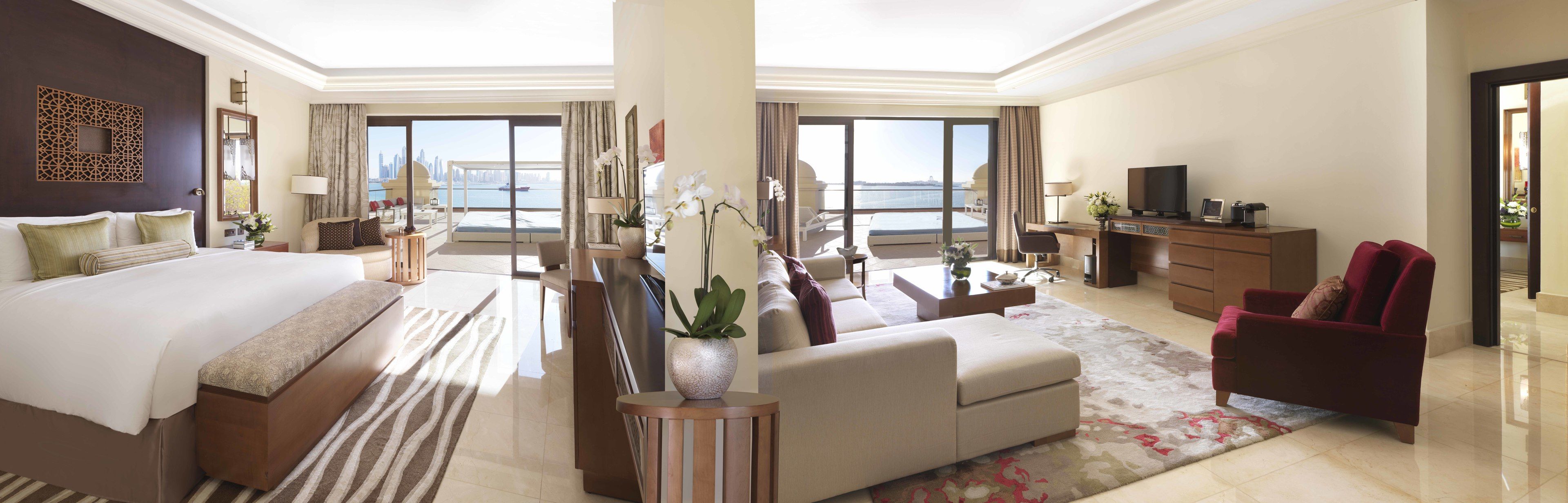A spacious hotel room with a large bed, a sofa, a desk, and a TV. The room has large windows with a view of the city skyline.