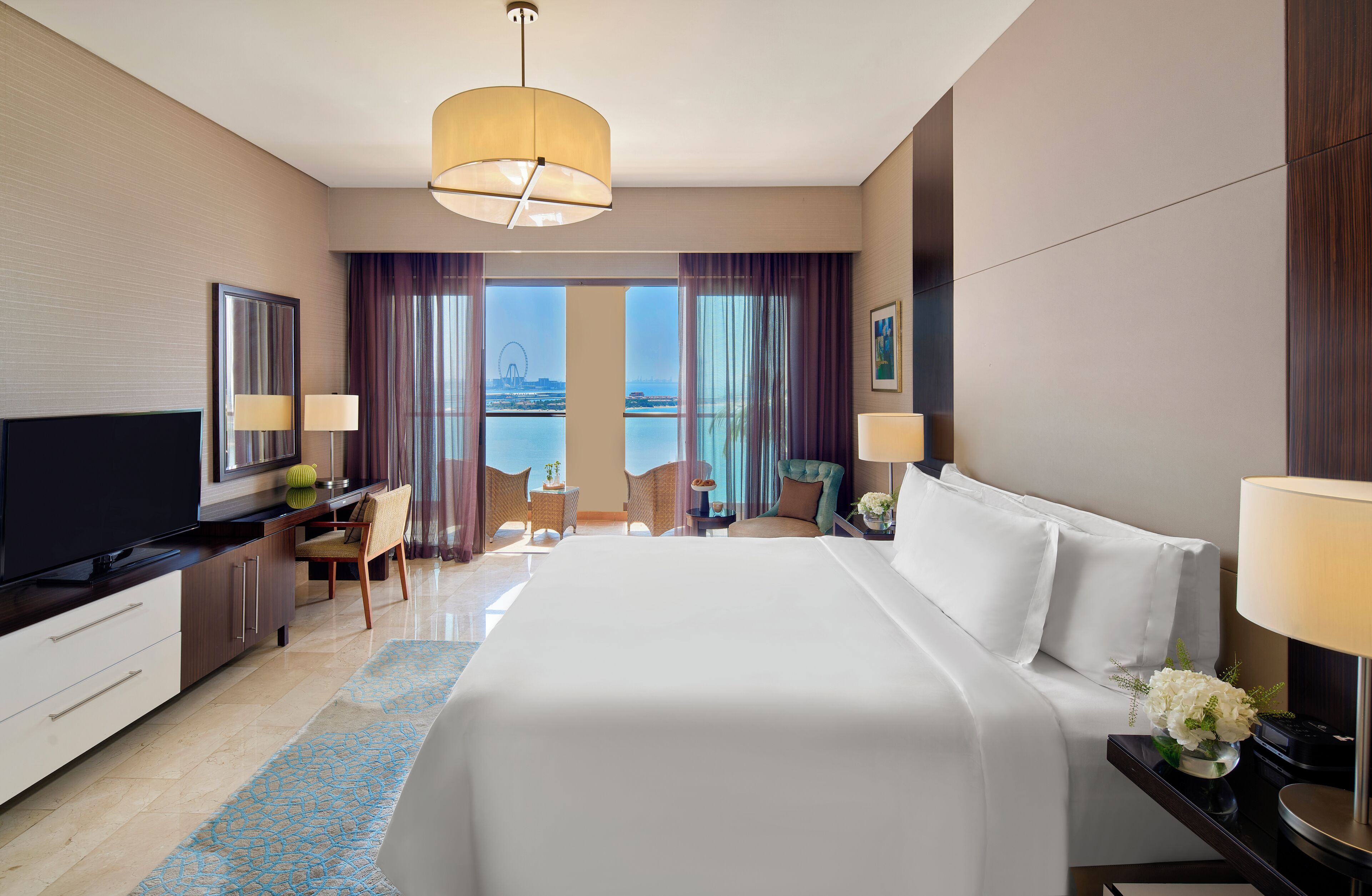 A modern hotel room with a large bed, a desk with a TV, a chair, and a view of the water.