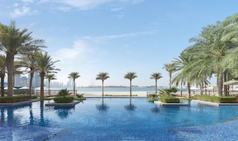 A swimming pool with clear blue water, surrounded by palm trees and a view of the sea and city skyline.