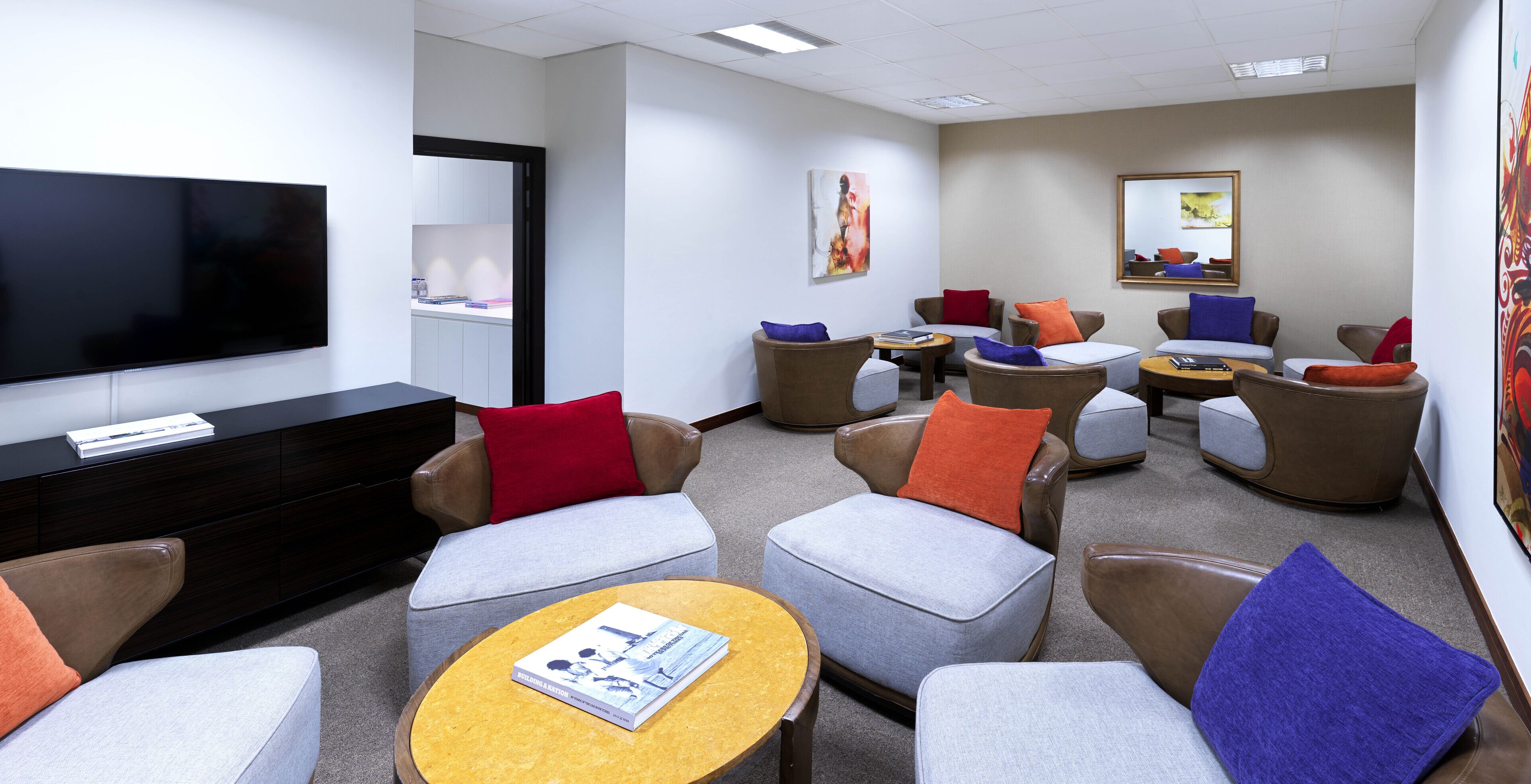 A hotel lobby with a flat-screen TV, multiple seating areas with armchairs and ottomans, a coffee table, and a bookshelf.