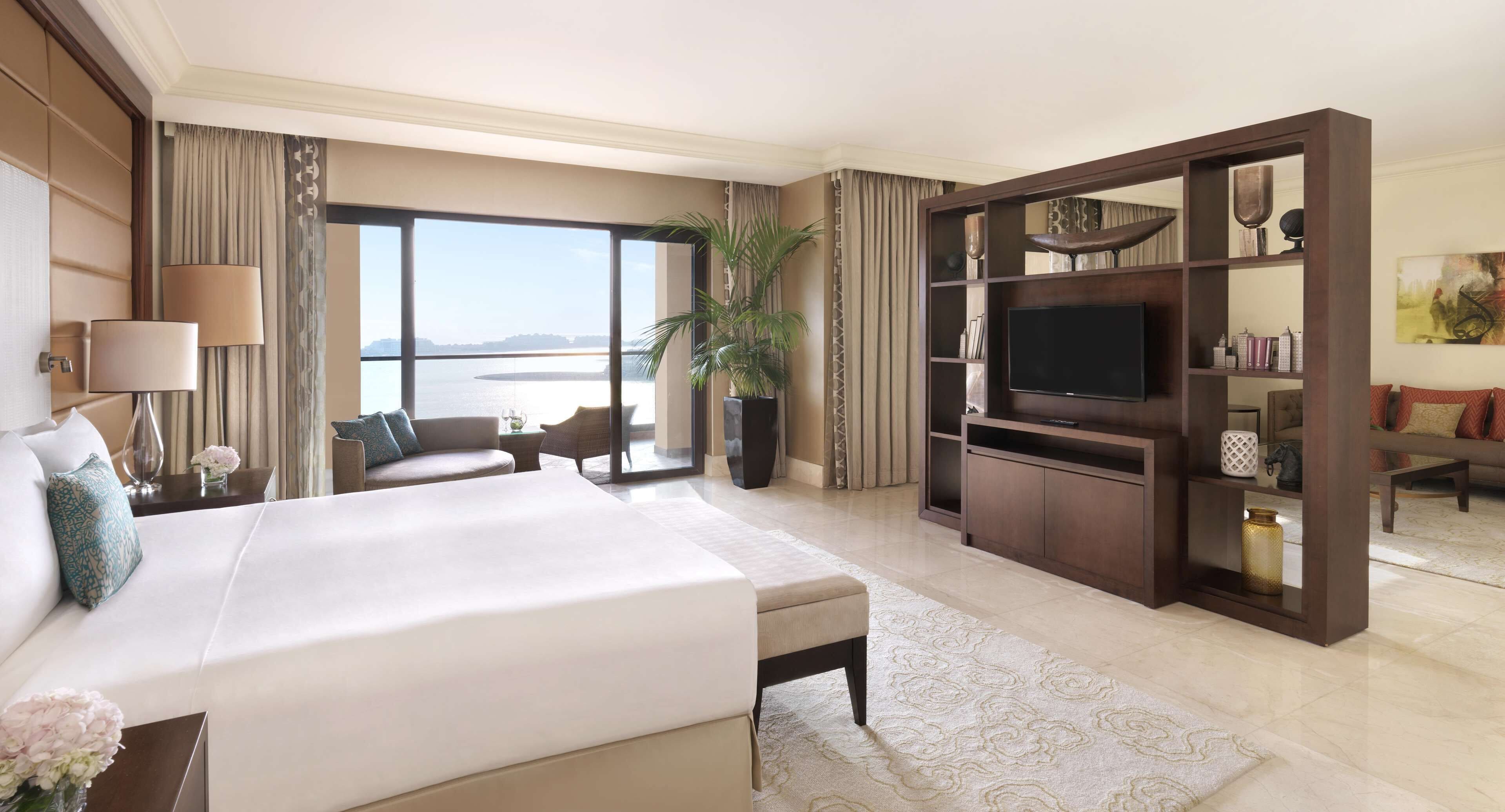 A hotel room with a large bed, a TV, a seating area with a view of the sea, and a bookshelf.