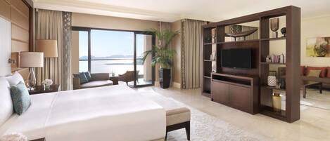 A hotel room with a large bed, a TV, a seating area with a view of the sea, and a bookshelf.