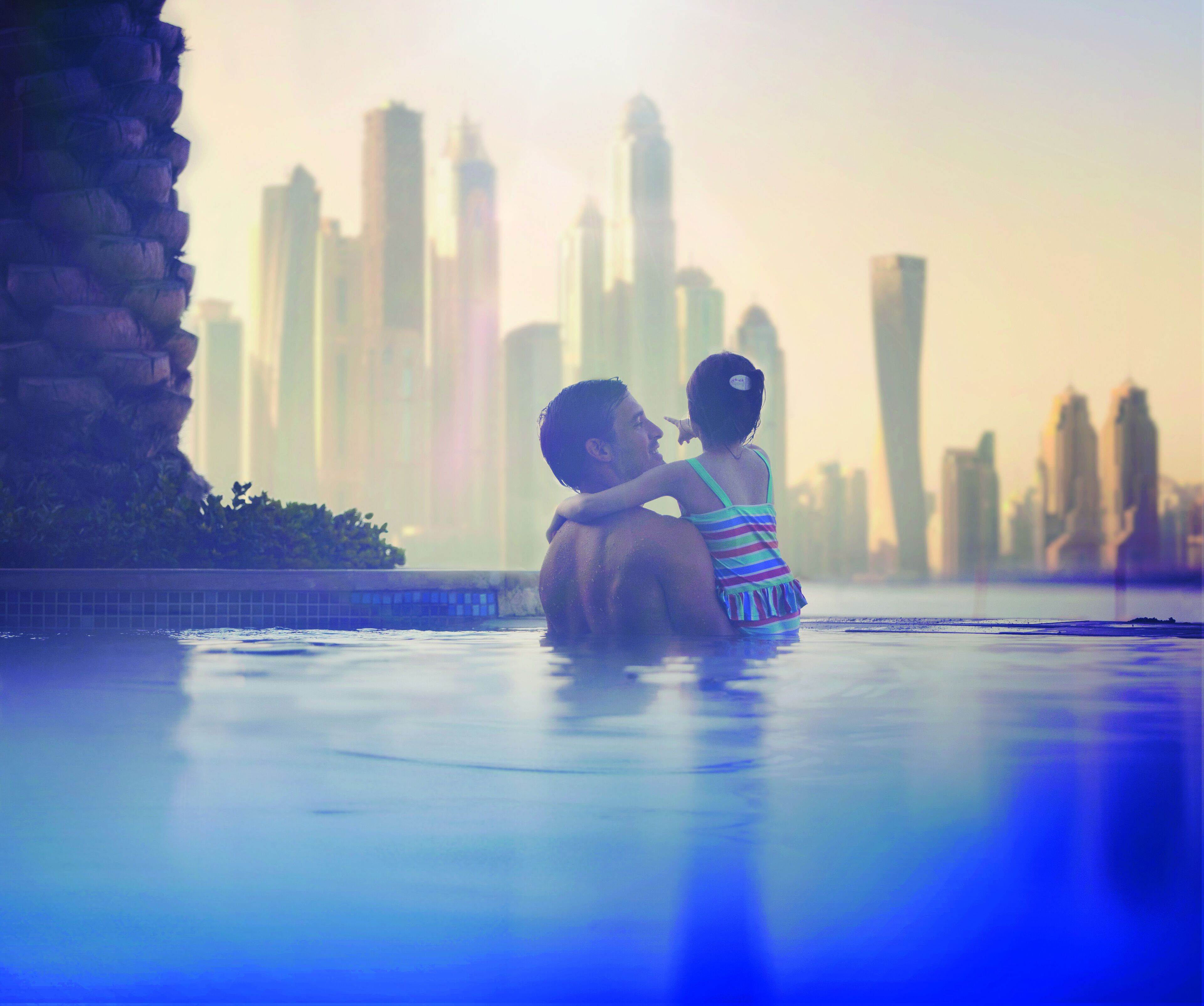 Una pareja se abraza en una piscina con un horizonte urbano al fondo.