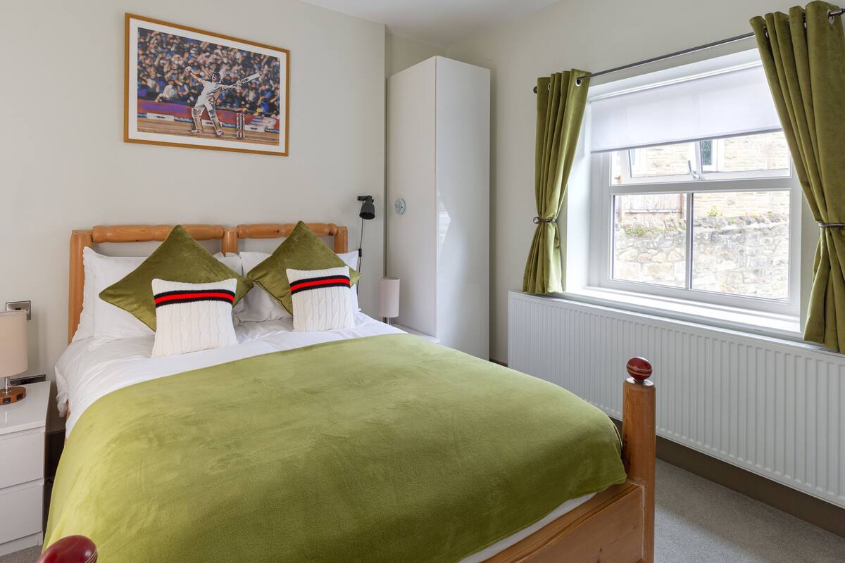 A neatly made bed with a green blanket, two pillows, and a wooden headboard. There is a window with green curtains and a radiator below it. A framed picture hangs on the wall above the bed.