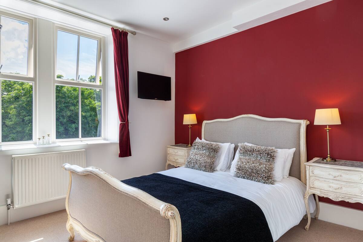 A bedroom with a large bed, two bedside tables with lamps, a TV on the wall, and a window with curtains.