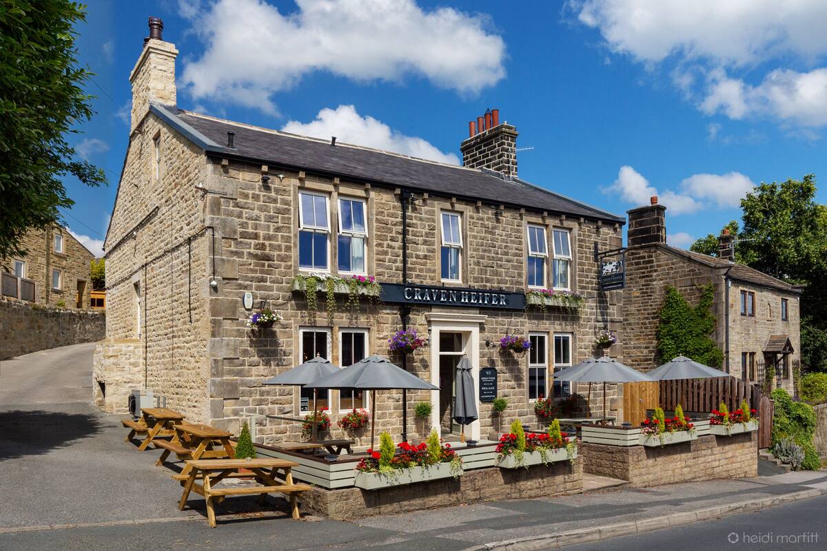 A stone building with a sign reading "Craven Heifer." It features outdoor seating with umbrellas and flower boxes.