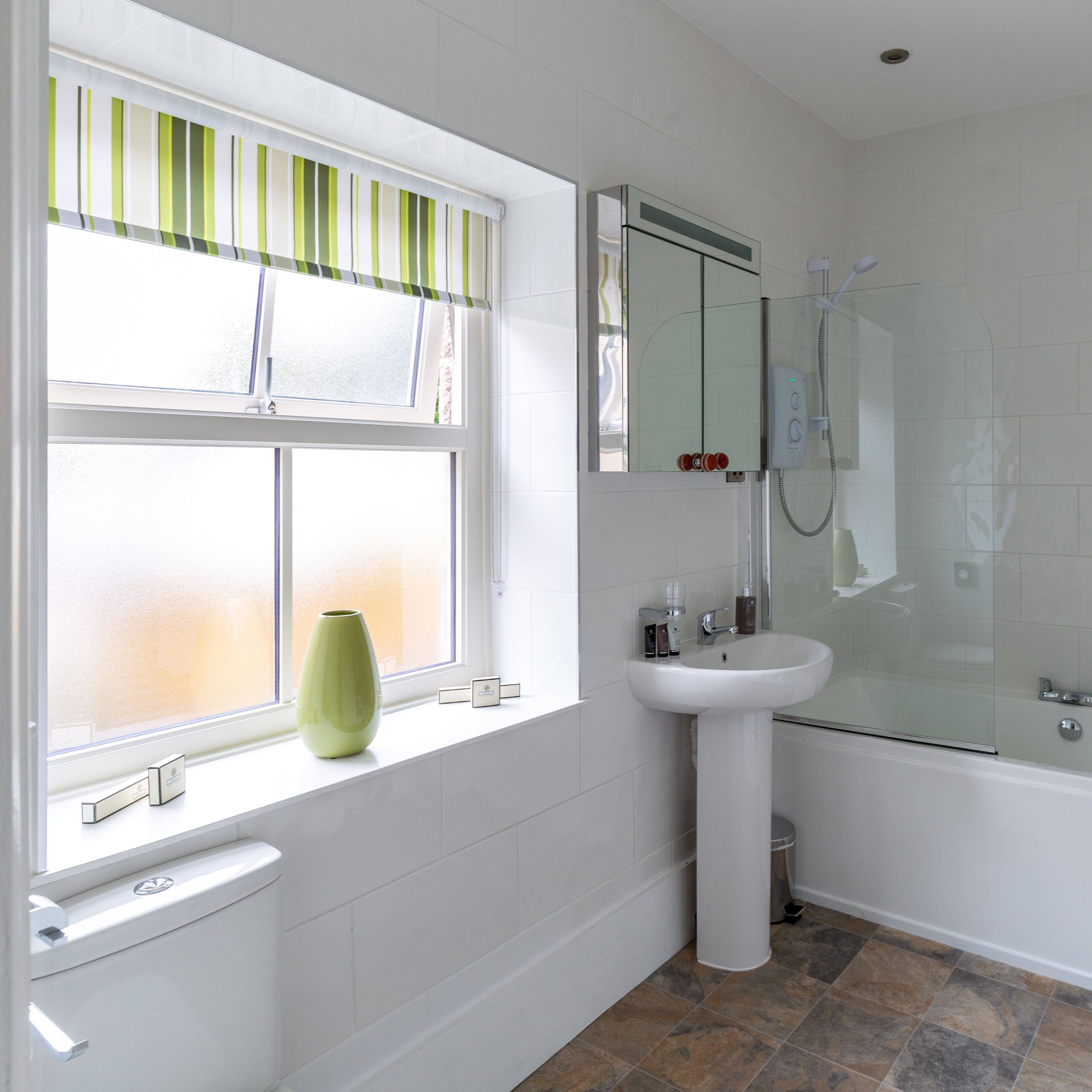 A bathroom with a toilet, a sink, a shower, and a window with a green-striped blind.
