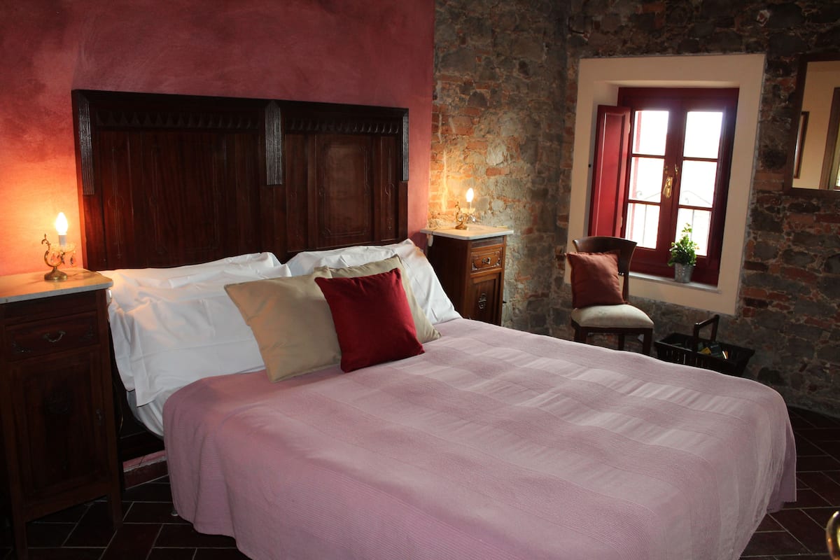 A bedroom with a large bed, wooden headboard, and a stone wall. There is a window with red shutters and a chair by the window.