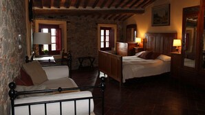 A rustic bedroom with a stone wall, wooden ceiling beams, a bed with a wooden headboard, a bedside table with a lamp, and a chair with a cushion. - San Gennaro Castello (Capannori)