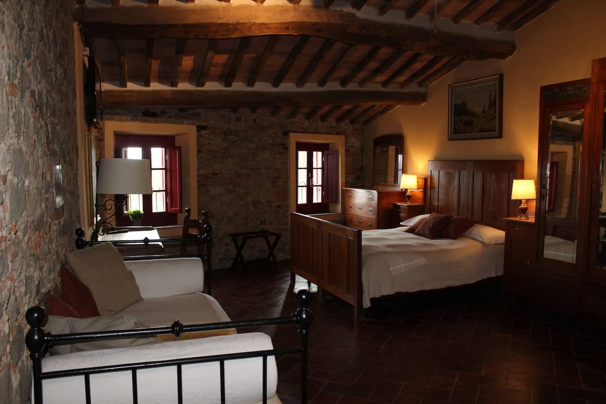 A rustic bedroom with a stone wall, wooden ceiling beams, a bed with a wooden headboard, a bedside table with a lamp, and a chair with a cushion.