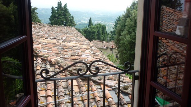 A view through a window with a metal railing, showcasing a terracotta-tiled roof and surrounding greenery. - San Gennaro Castello (Capannori)