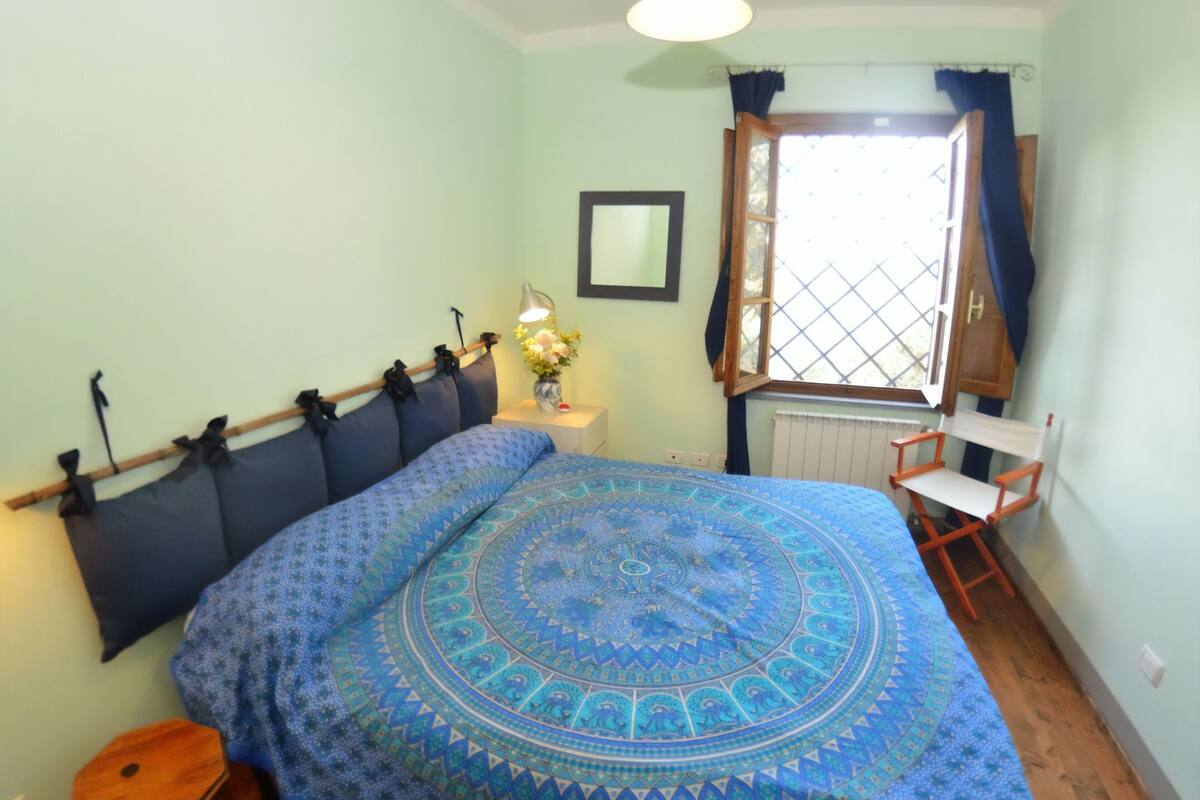 A bedroom with a bed, a nightstand, a chair, a window with curtains, and a radiator.