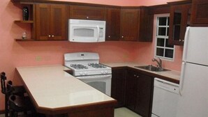 A compact kitchen with a white stove, microwave, and refrigerator, wooden cabinets, and a small window. - Piarco Village Suites (Piarco)
