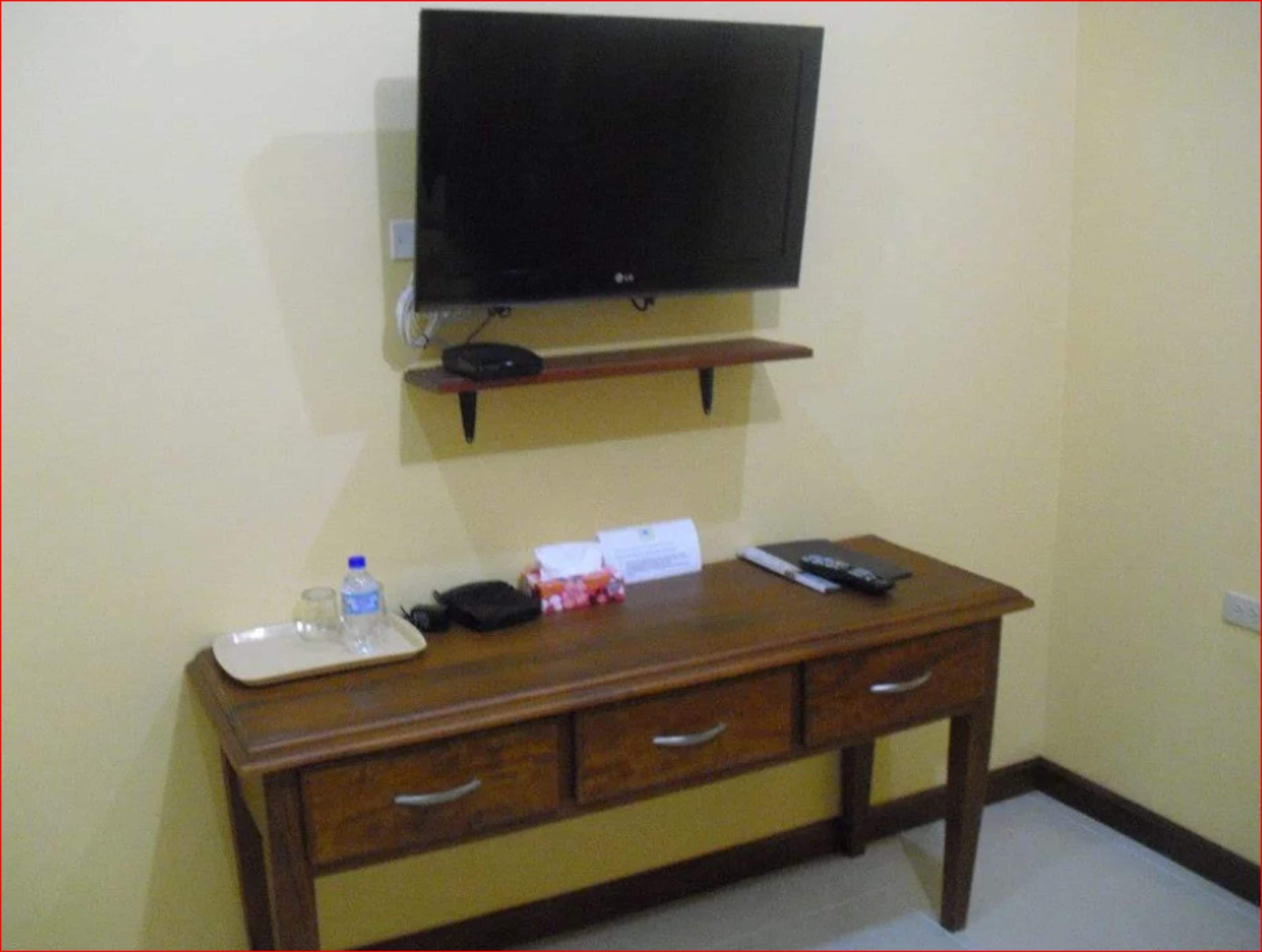 A hotel room with a wooden desk, a television mounted on the wall, a water bottle, a remote control, and a small box.