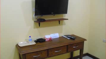 A hotel room with a wooden desk, a television mounted on the wall, a water bottle, a remote control, and a small box.