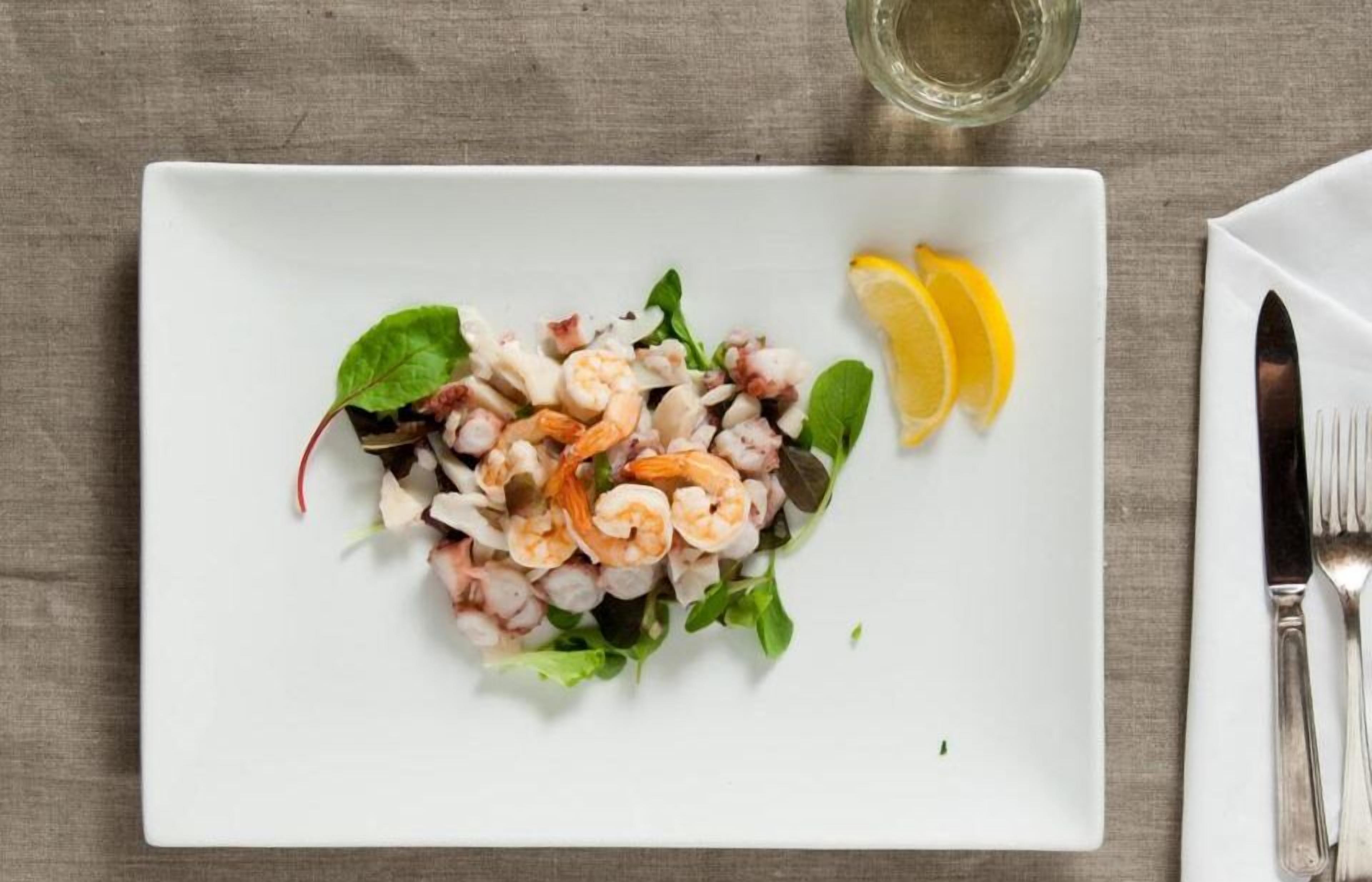 Ein weißer Teller mit einem Seafood-Salat, Zitronenspalten und einem Glas Wasser auf einem Tisch.