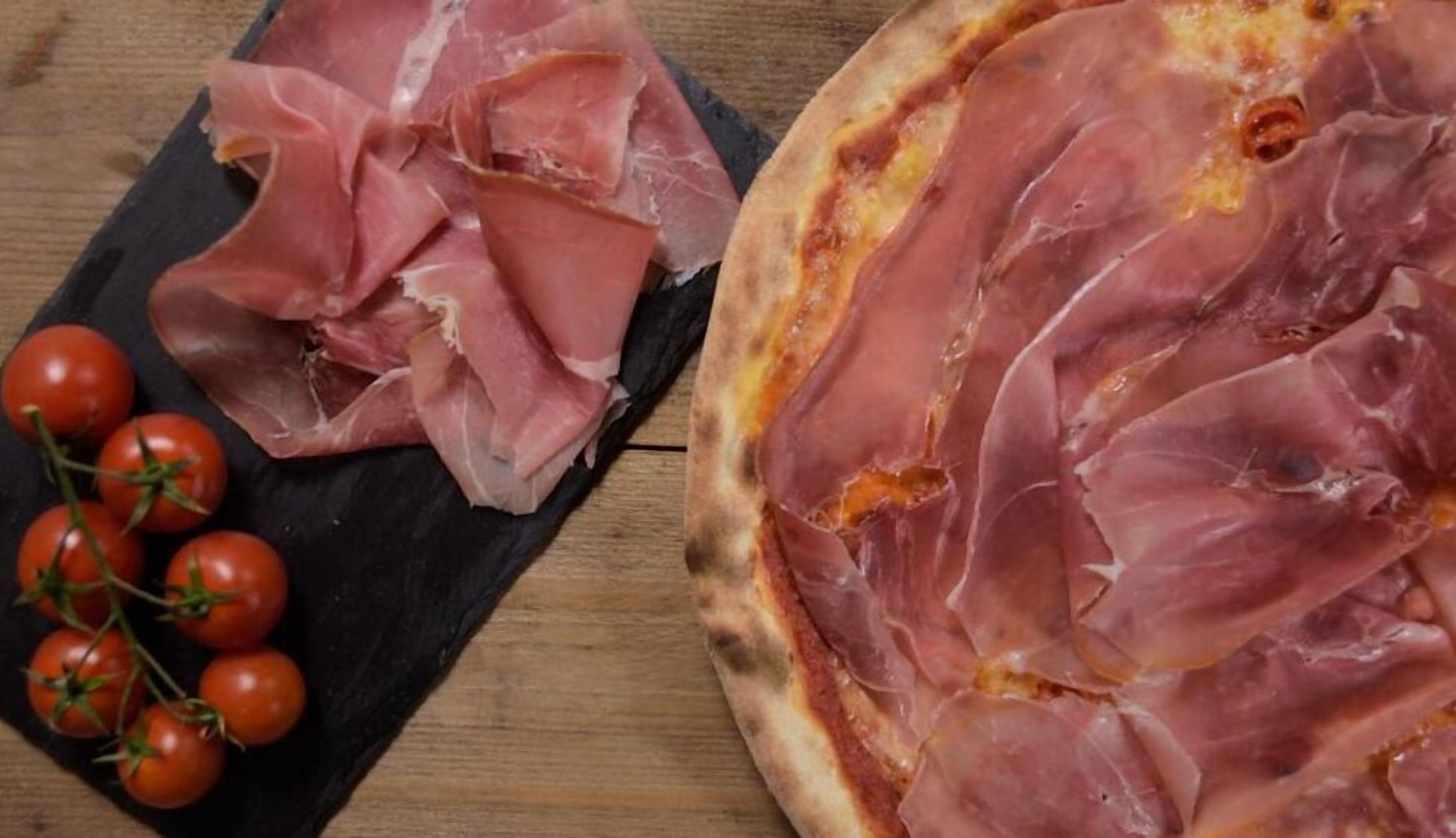 A pizza with prosciutto slices on a black slate board, accompanied by cherry tomatoes.
