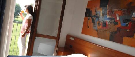 A hotel room with a bed, a wooden headboard, a large abstract painting, and a balcony with a view of greenery.