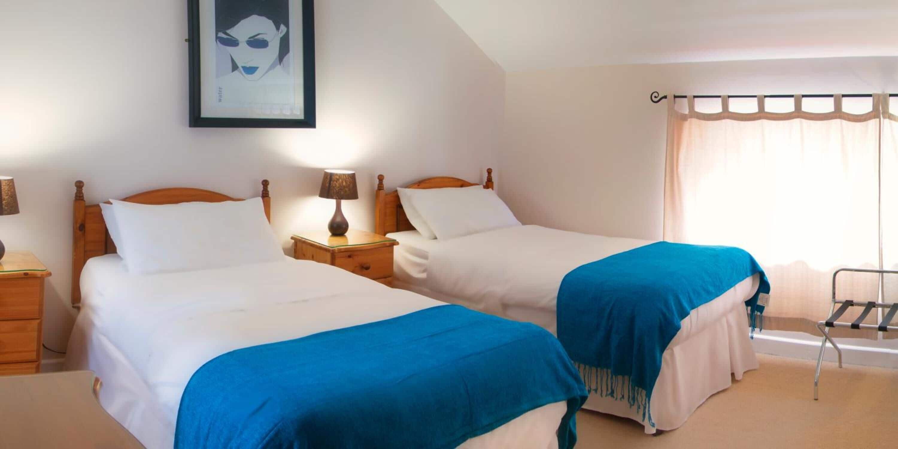 A hotel room with two single beds, a nightstand with a lamp, a framed picture on the wall, and a window with a curtain.