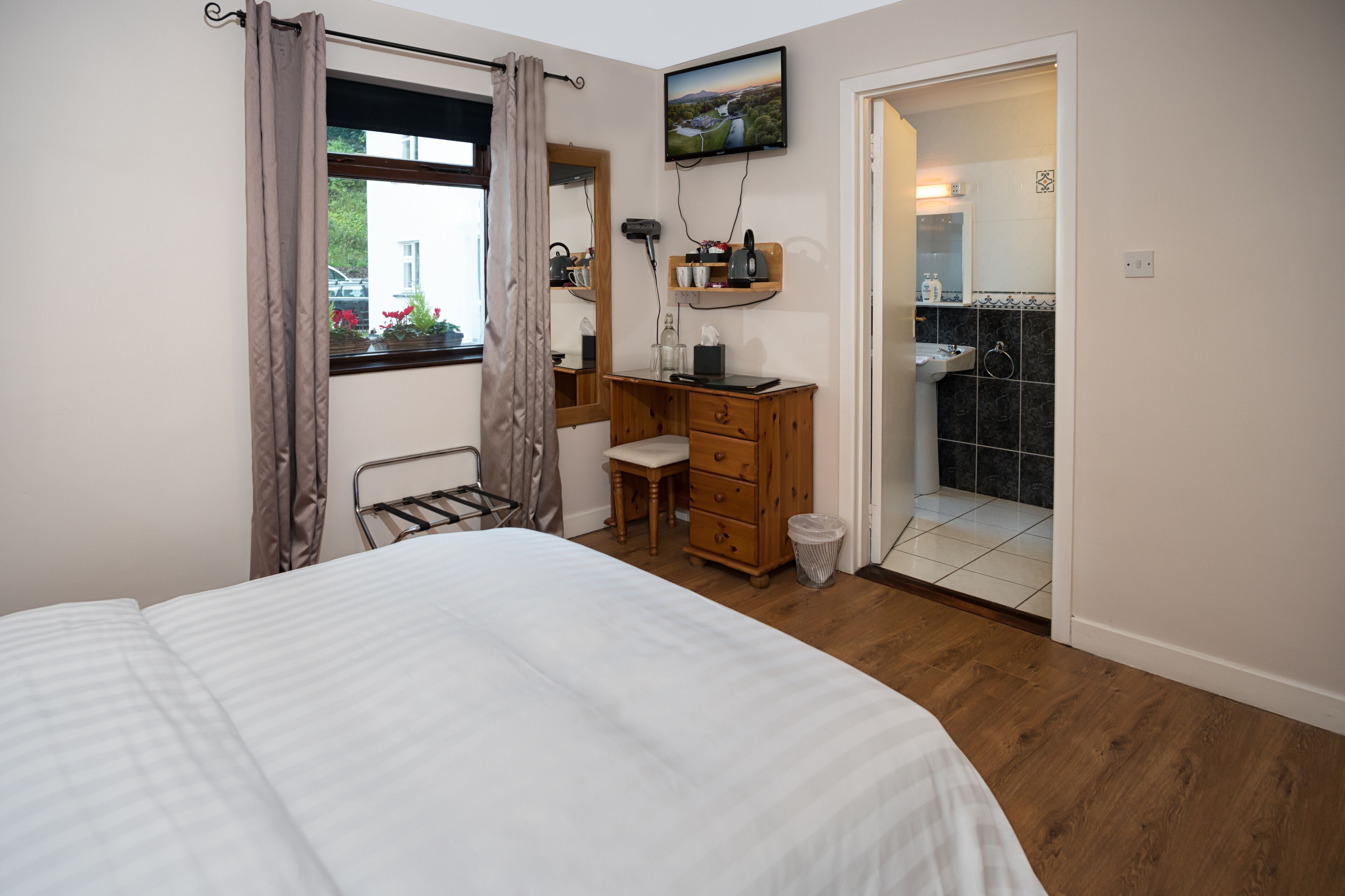 A bedroom with a bed, a wooden dresser, a desk, a mirror, a TV, and a bathroom visible through an open door.