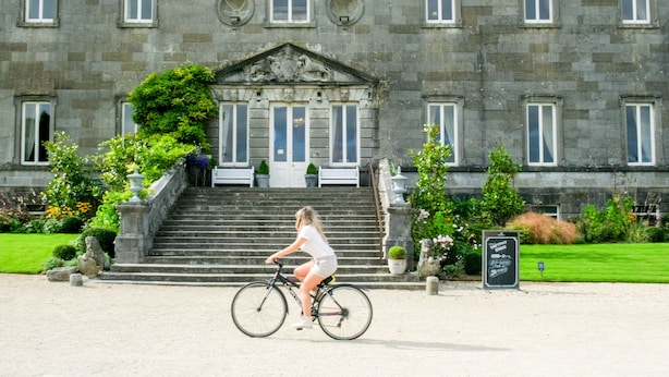 A woman riding a bicycle in front of a stone building with a grand entrance, steps, and a signboard. - Woodside Lodge (Westport)