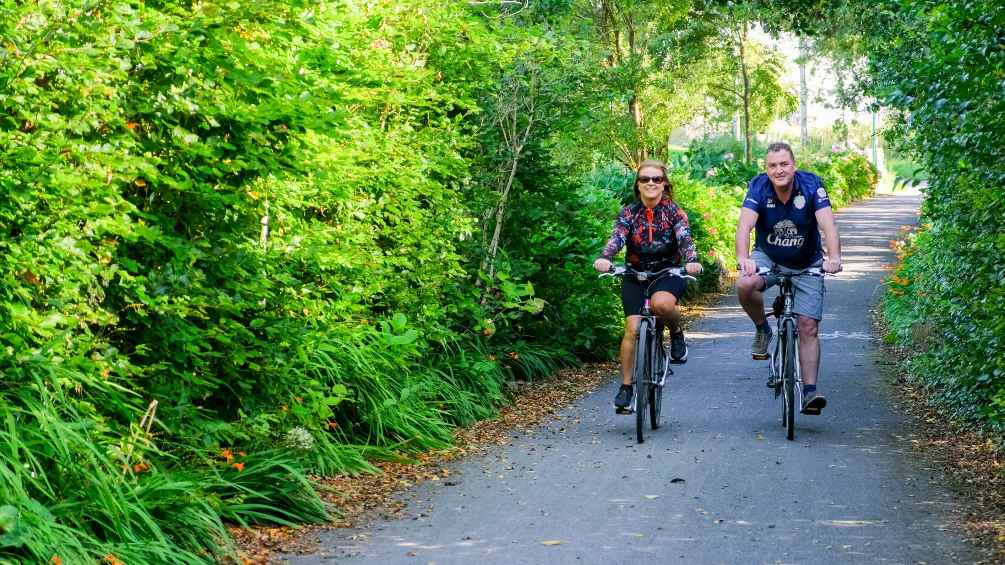 Two cyclists on a paved path surrounded by dense green foliage.