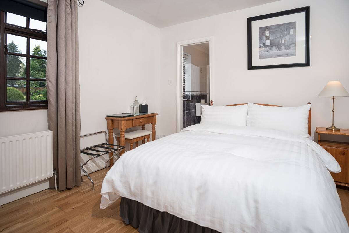 A bedroom with a bed, a wooden nightstand, a lamp, a radiator, a window with curtains, and a framed picture on the wall.