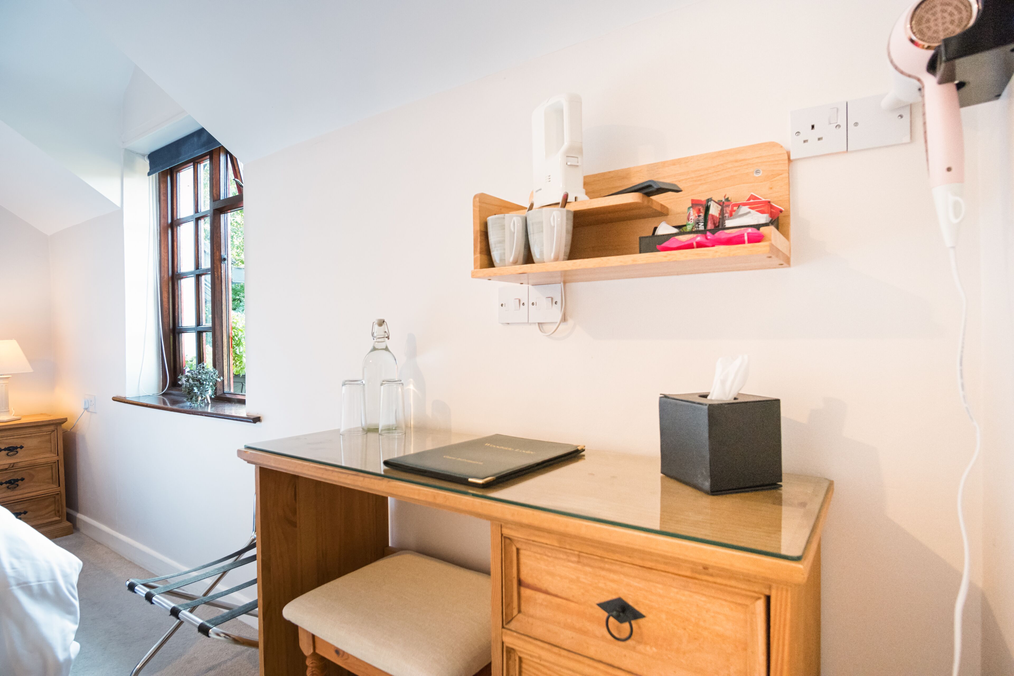 Habitación con escritorio de madera, ventana con vistas, estante con artículos de aseo e un secador de pelo enchufado.