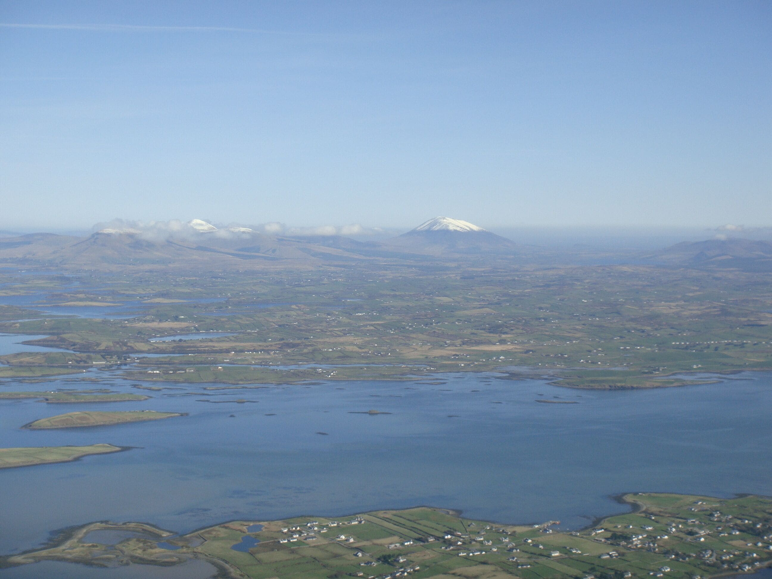 An aerial view of a coastal area with a mountain range in the background, water bodies, and scattered settlements.
