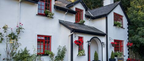 A white building with red window frames and black trim, surrounded by greenery and potted plants.
