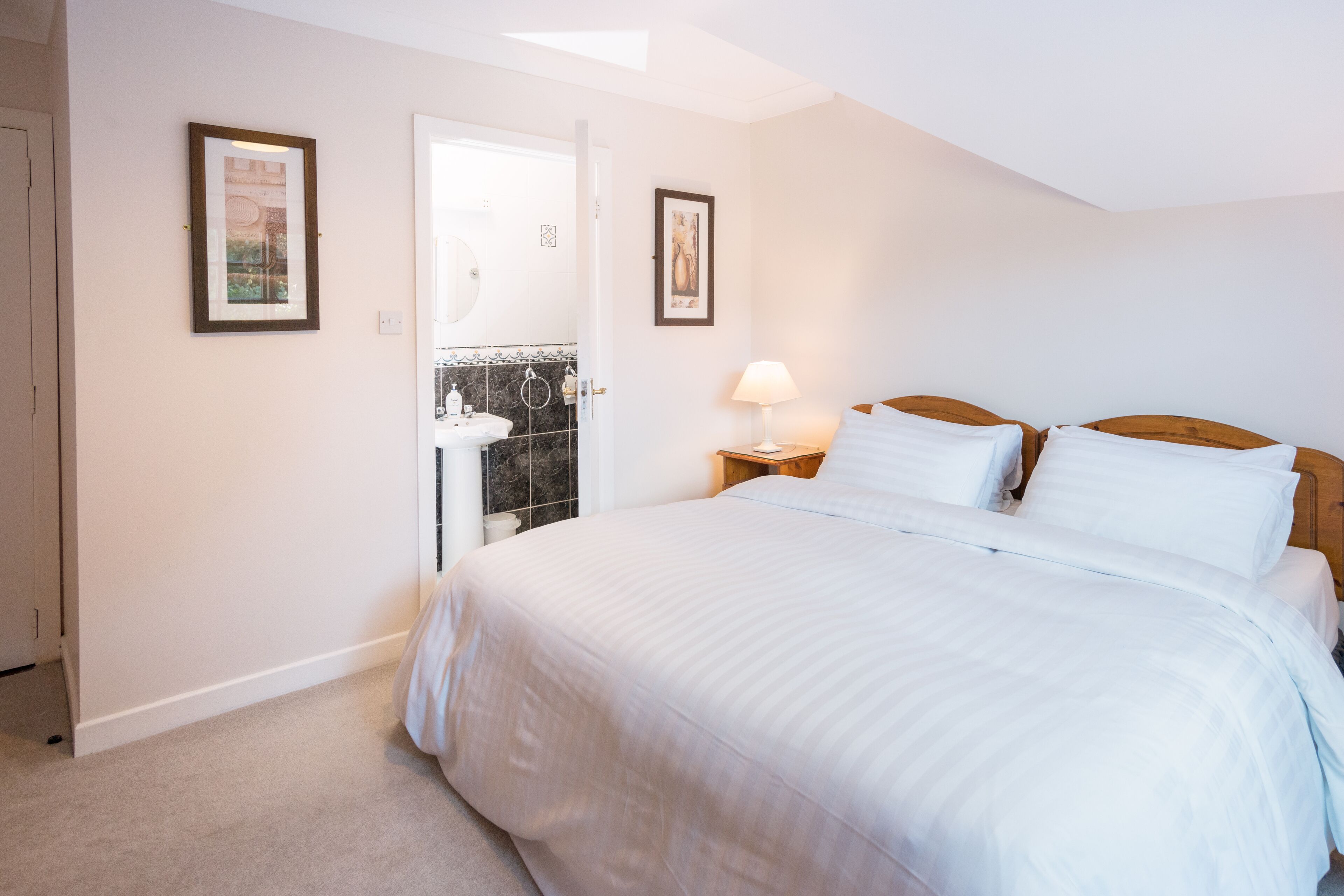 Une chambre à coucher avec un grand lit, une table de chevet avec une lampe, et une salle de bain visible à travers une porte ouverte.