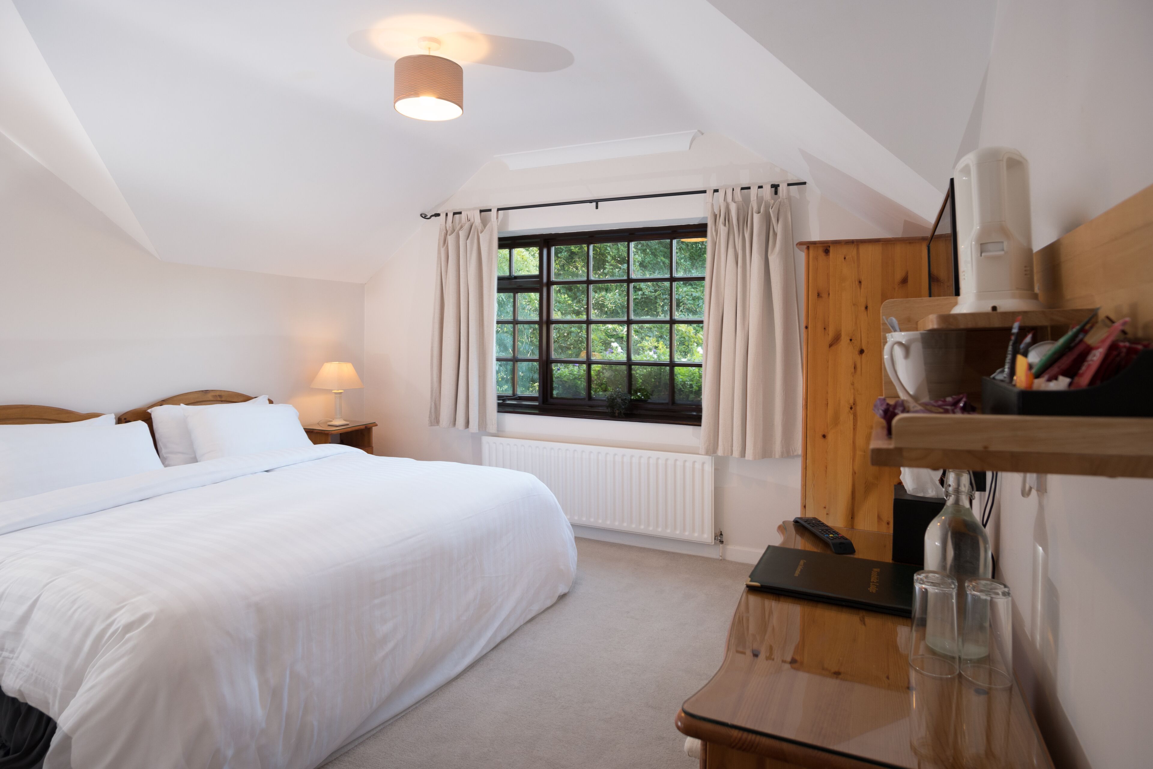 A bedroom with a large bed, a window with curtains, a bedside table with a lamp, and a wooden shelf with items.
