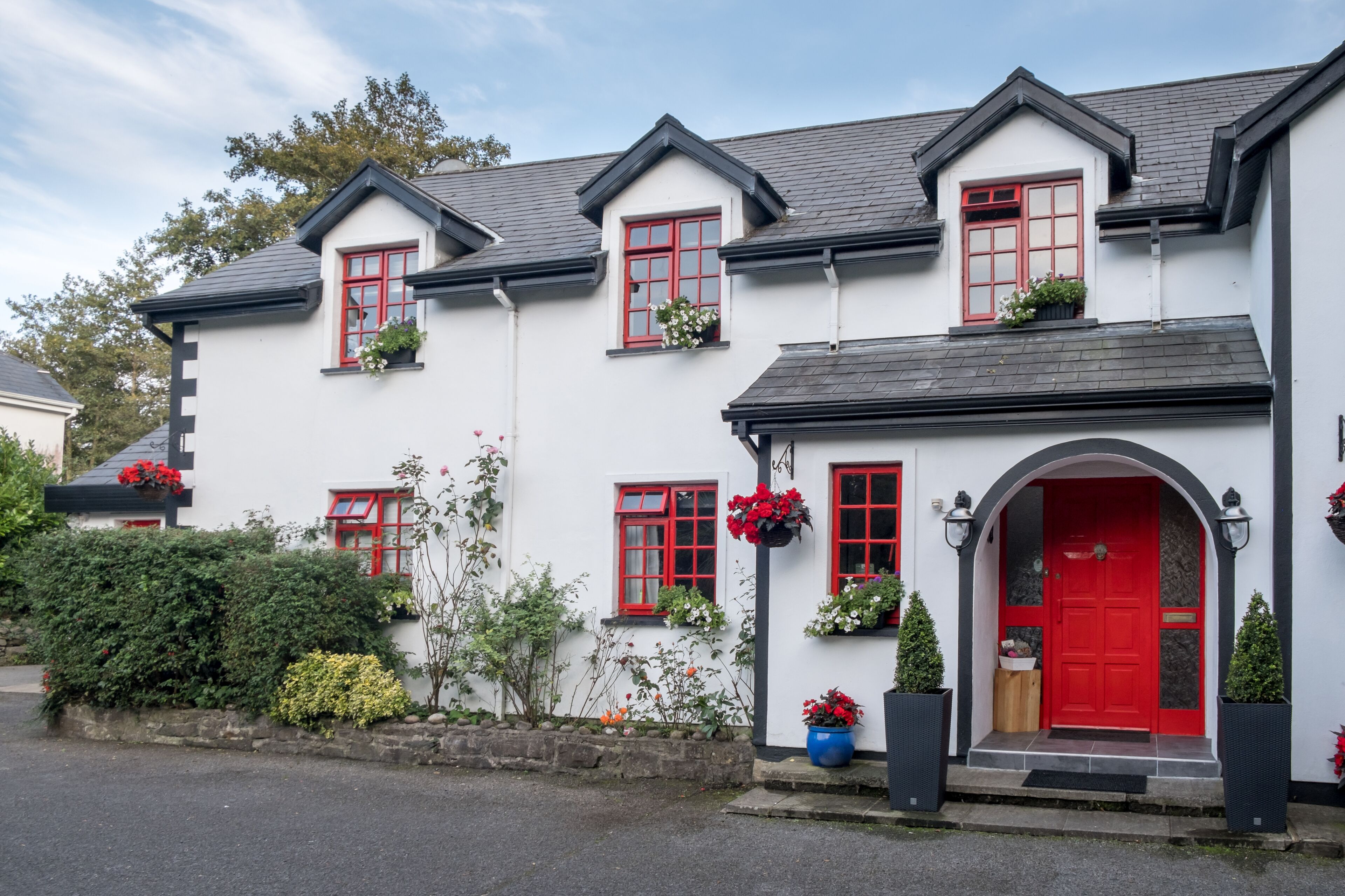 A white building with red windows and a red door, surrounded by greenery and potted plants.