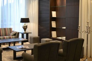 A modern living room with a sofa, armchairs, a coffee table, and a lamp. There is a bookshelf with books and a decorative item. - Blue Woods Hotel (Deerlijk)