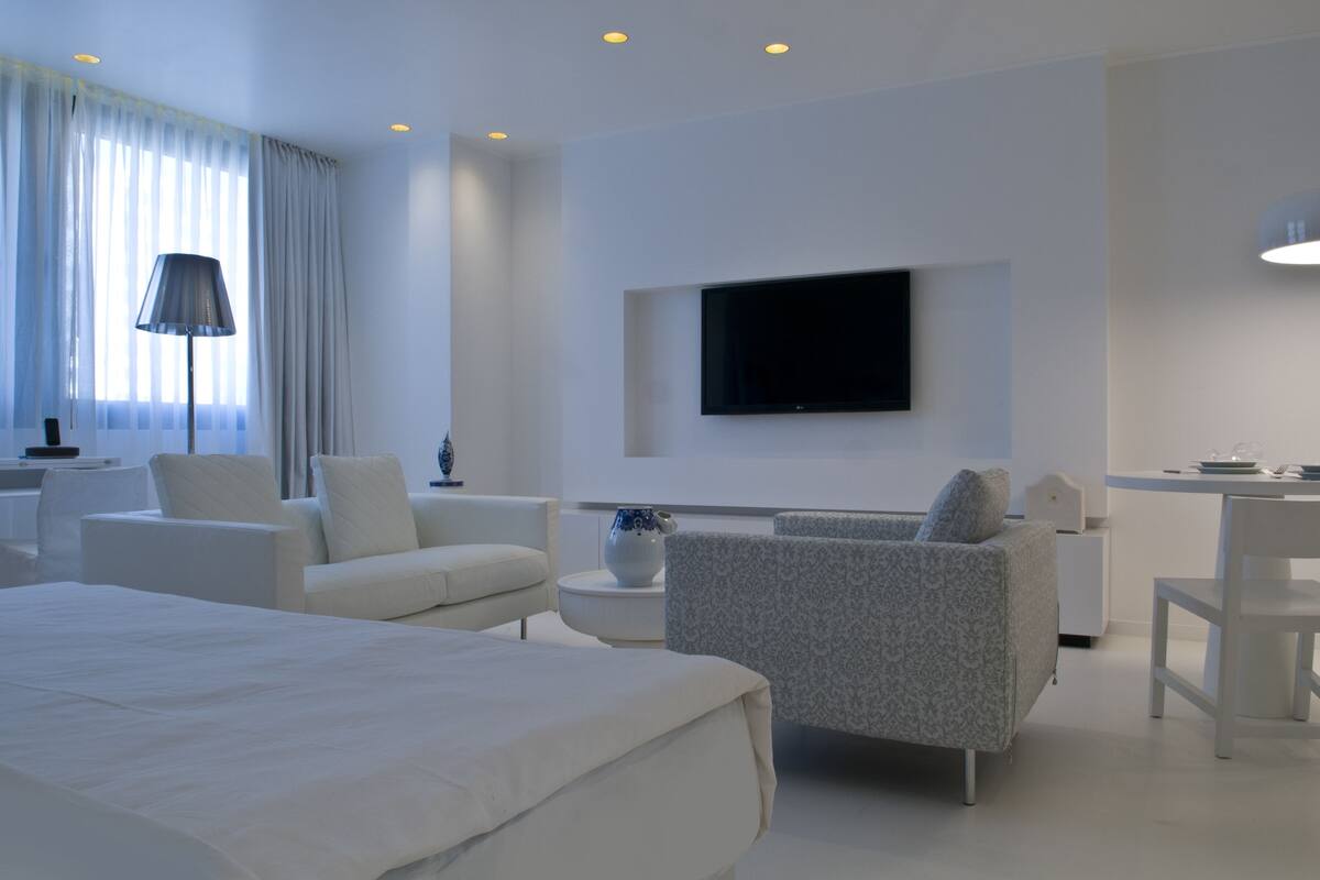 Una habitación de hotel moderna con una cama grande, dos sillones, una mesita, un televisor montado en la pared y un escritorio con una silla.