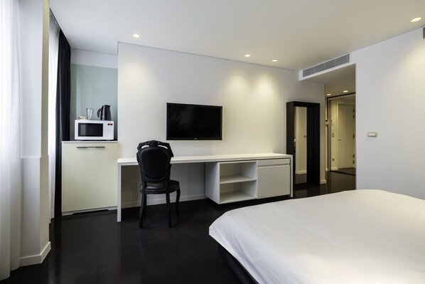A modern hotel room with a large bed, a desk with a chair, a flat-screen TV, a microwave, and a small kitchenette.