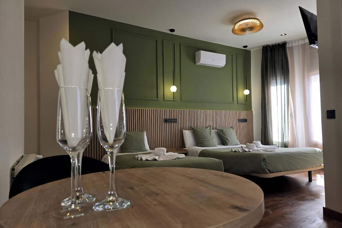 A hotel room with a bed, bedside tables, a round wooden table with two glasses, a wall-mounted air conditioner, and a ceiling light fixture.