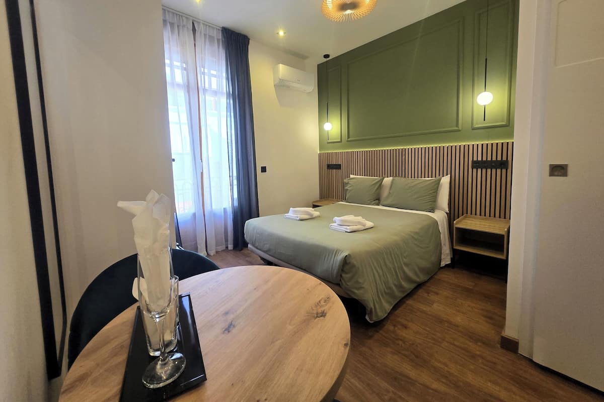 A hotel room with a bed, bedside table, and a dining table with a glass.