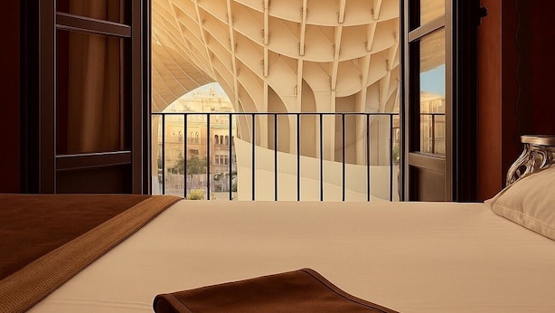 Habitación de hotel con una cama grande, vistas de un edificio a través de un muro de cristal y un balcón con un diseño de techo singular.