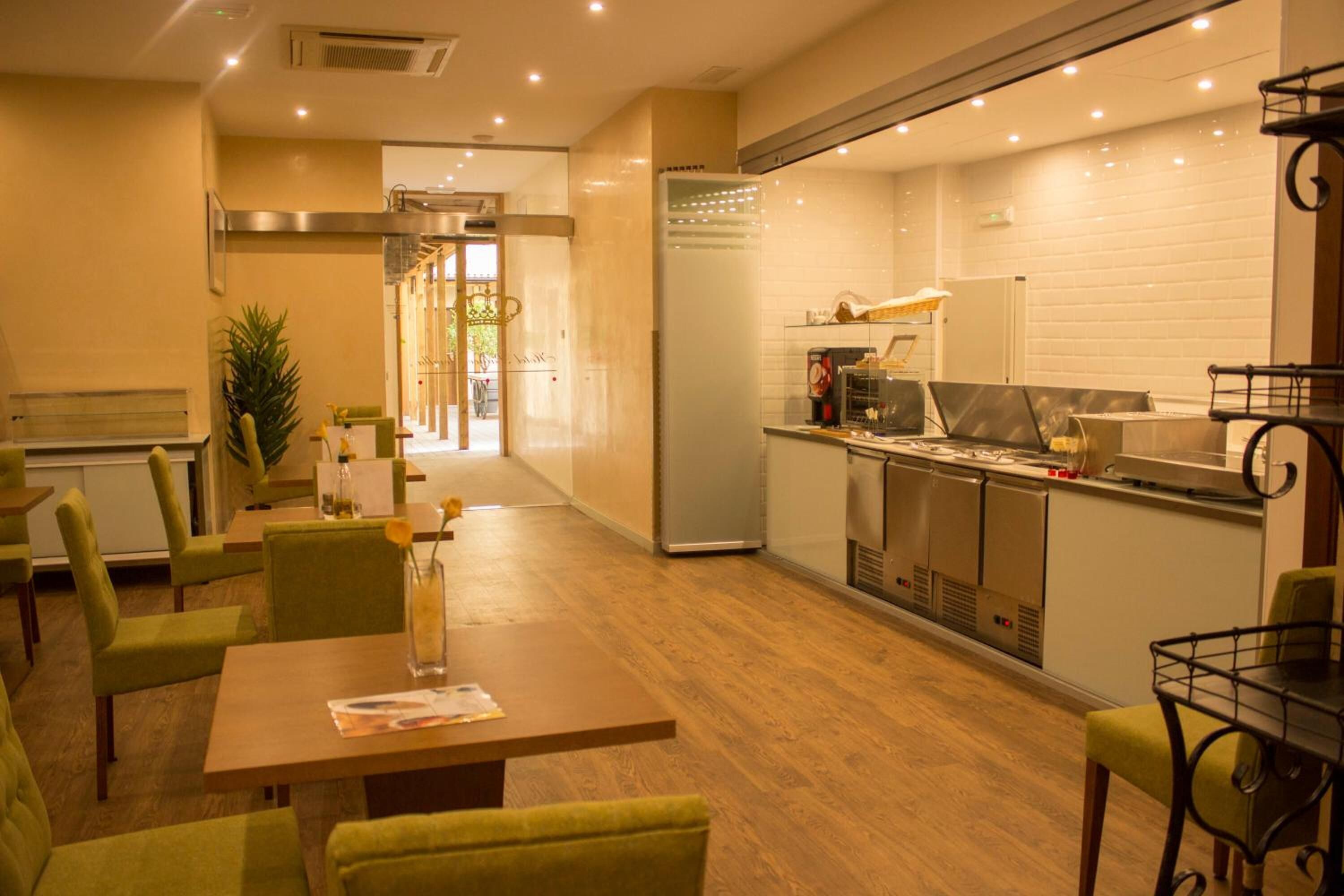 Um restaurante com piso de madeira, cadeiras estofadas em verde, uma mesa de jantar central e uma área de cozinha com eletrodomésticos de aço inoxidável.