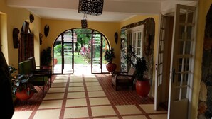 Un pasillo amplio con suelo de baldosas, una silla verde, una maceta roja y una puerta que da al exterior.