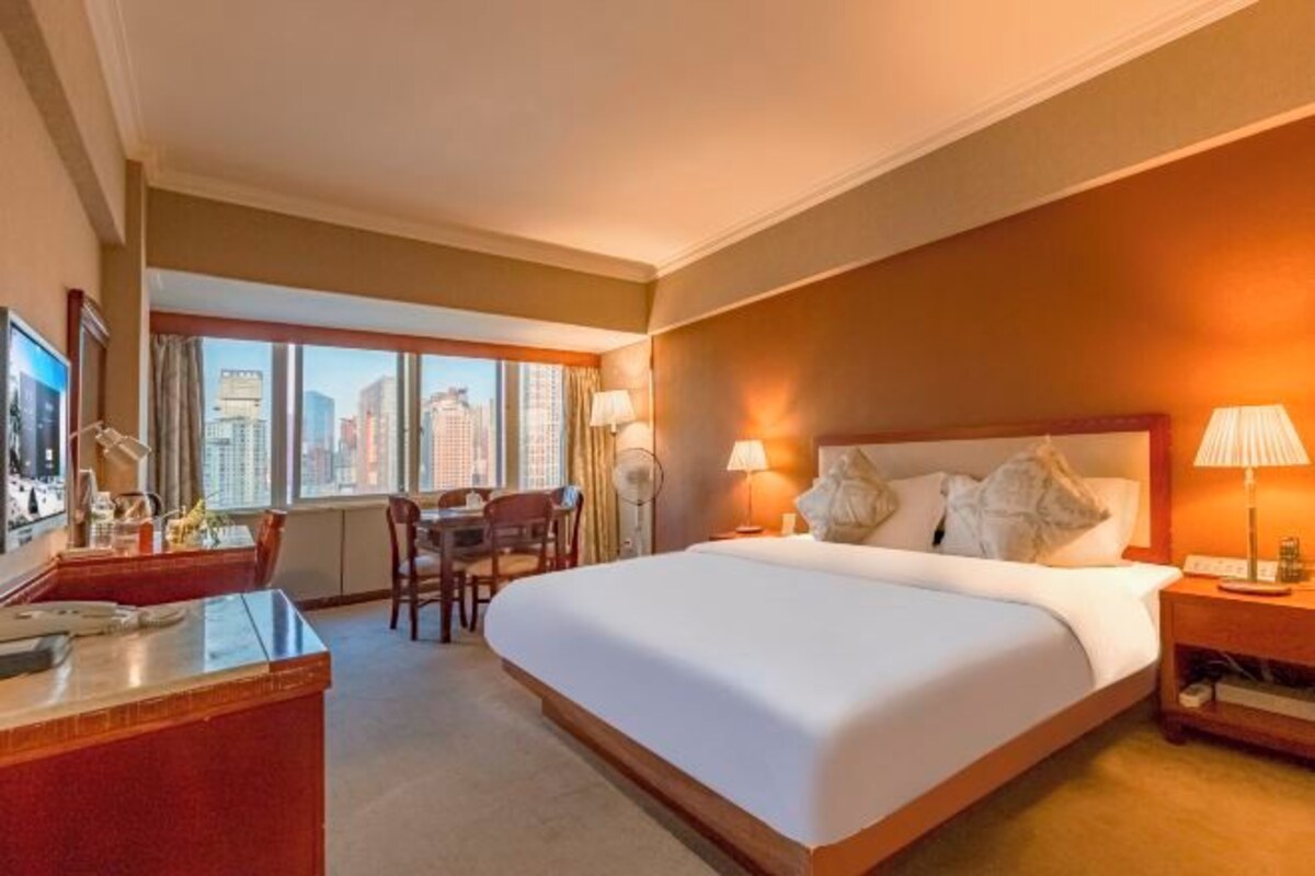 A hotel room with a large bed, a desk, a chair, and a view of the city through large windows.
