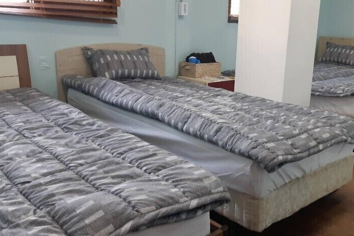 Une chambre de dortoir avec trois lits, chacun recouvert d’une couette et de coussins à motifs gris. On y trouve une tête de lit en bois sur le côté gauche, une petite table avec une boîte, et un pilier blanc au centre.