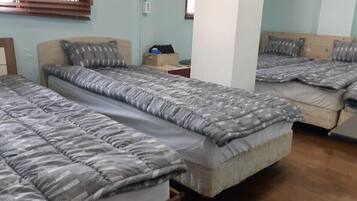 Habitación tipo dormitorio con tres camas, cada una con una colcha y almohadas grises de estampado. Se observa un cabecero de madera a la izquierda, una mesita con una caja y un pilar blanco en el centro.